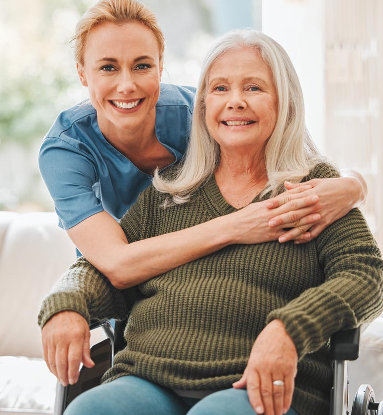 senior-healthcare A smiling caregiver embraces a senior woman in a wheelchair inside a cozy home setting.