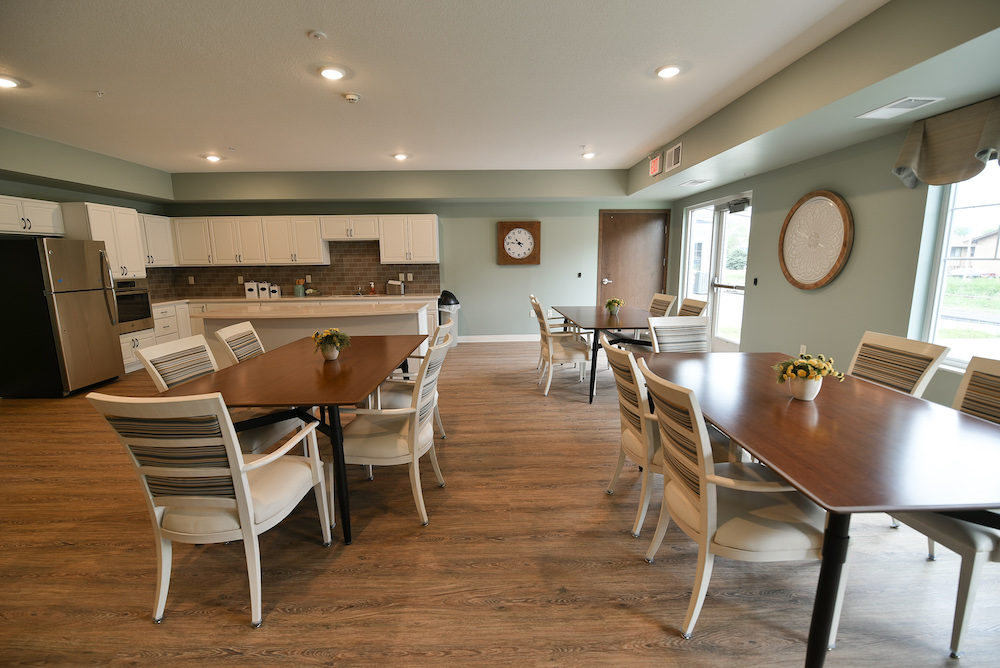 A well-lit communal dining area with wooden tables, white chairs, and a kitchen in the background.