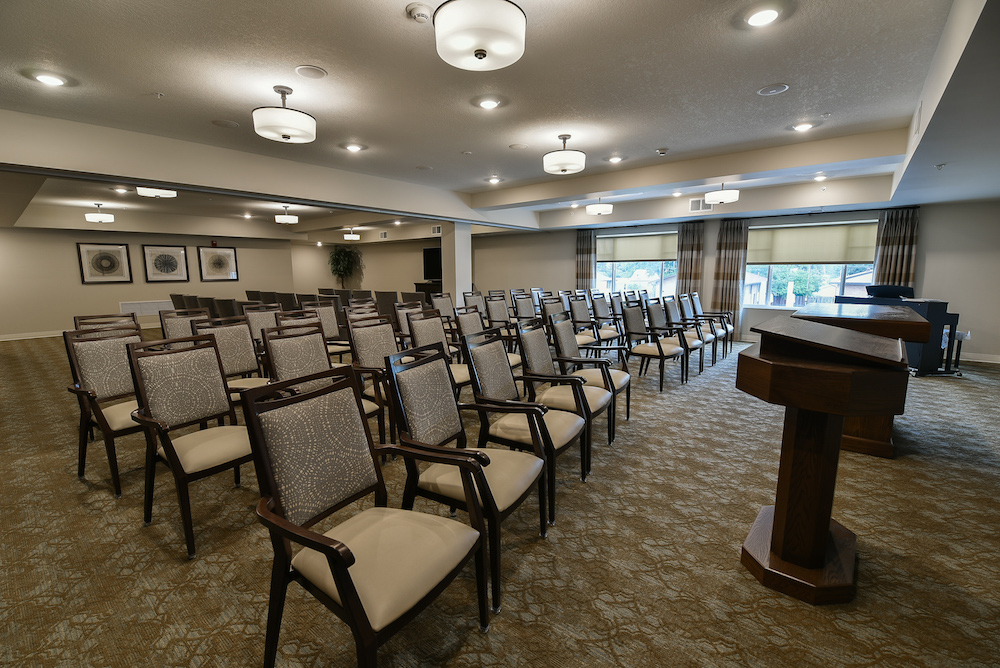 An empty conference room with rows of chairs facing a wooden podium and large windows in the background.