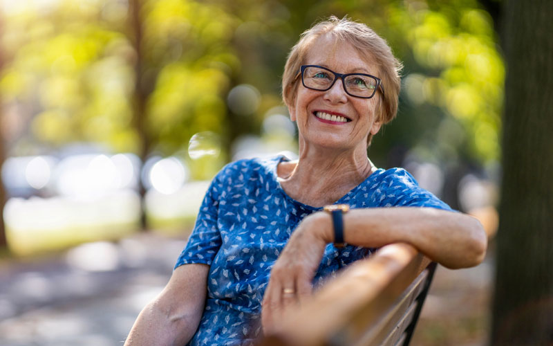 smiling-senior Happy senior woman with glasses smiling while sitting on a park bench on a sunny day.